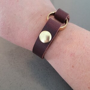 May include: A brown leather bracelet with a gold metal ring and a gold button closure.