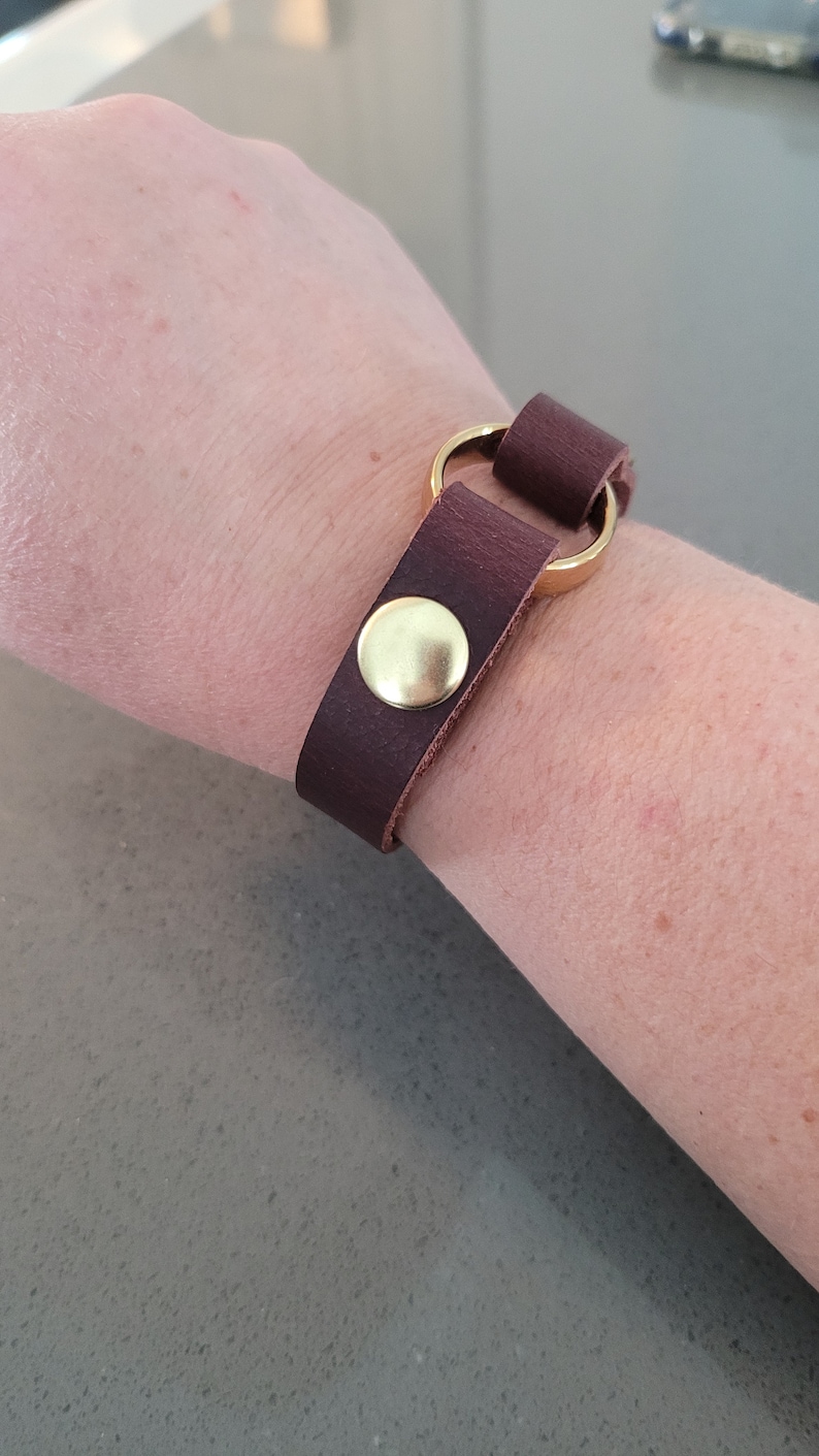 May include: A brown leather bracelet with a gold metal ring and a gold button closure.