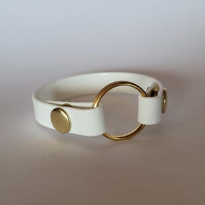 Peut inclure: Un bracelet blanc avec une finition lisse et brillante, agrémenté d'un anneau central en métal doré. La bande est fixée par deux boutons-pression en métal doré, placés de chaque côté de l'anneau, formant un design circulaire épuré.