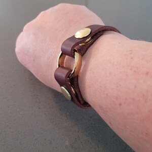 May include: A brown leather bracelet with gold metal accents. The bracelet has a circular design and is fastened with a buckle.