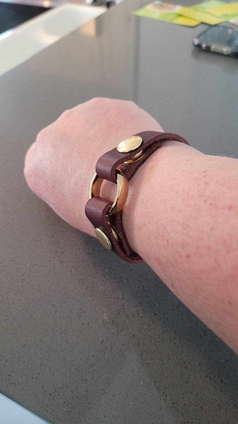 May include: A brown leather bracelet with gold metal accents. The bracelet has a circular design and is fastened with a buckle.