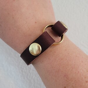May include: A brown leather bracelet with a gold metal ring and two gold metal snaps.