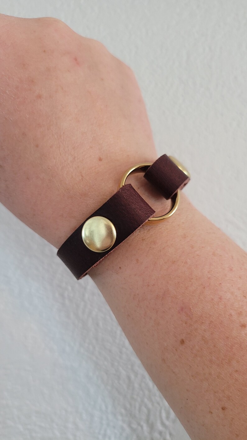 May include: A brown leather bracelet with a gold metal ring and two gold metal snaps.