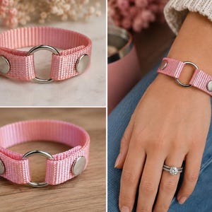 Peut inclure: Un bracelet en sangle de nylon rose clair doté d'un anneau central en métal argenté et de deux boutons-pression en métal argenté. La sangle est fabriquée dans un tissu tissé durable, l'anneau métallique servant de point focal entre les deux extrémités.
