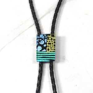 May include: A black bolo tie with a rectangular glass pendant. The pendant has a geometric pattern in blue, green, and gold.