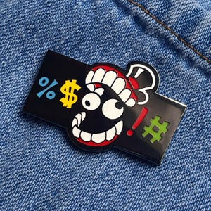 May include: Black enamel pin with a cartoon face design. The pin features a smiling face with red lips, white teeth, and a red and white hat. Symbols include a percent sign, dollar sign, exclamation point, and a hashtag.
