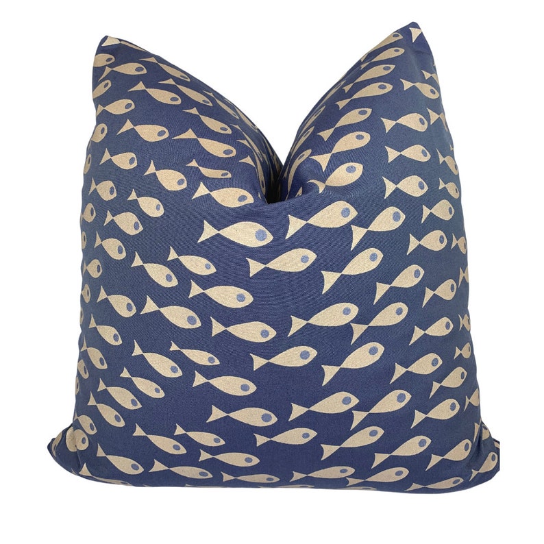 May include: A square cushion featuring a navy blue fabric adorned with a repeating pattern of beige fish. The fish design is stylized and arranged in a flowing pattern. The cushion has a soft appearance, suitable for home decor.