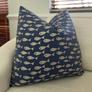 May include: Square navy blue cushion featuring a repeating pattern of beige fish. The cushion is made of a soft fabric and is placed on a light-coloured sofa. Ideal for adding a nautical touch to a living space.