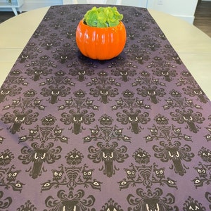 May include: A purple table runner with a repeating pattern of a haunted house with ghosts and owls. The pattern is in black and white. A small orange pumpkin with a green plant inside is sitting on the table runner.