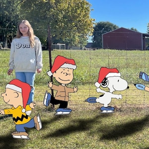 May include: A group of Peanuts characters, including Charlie Brown and Snoopy, ice skating in a grassy field, each wearing a Santa hat. A person wearing a gray sweatshirt stands in the background. The sky is blue.