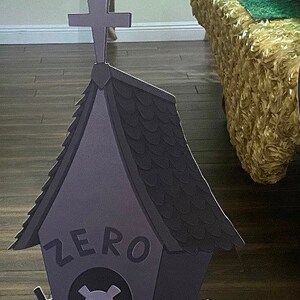 May include: A black and grey cardboard cutout of a dog house with a cross on top. The dog house has a sign that says "ZERO" and a skull and crossbones inside. The dog house is on a wooden floor.