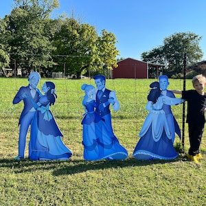 May include: A young boy stands in front of a fence with four blue cardboard cutouts of couples dancing. The cutouts are dressed in 19th-century attire.