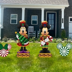 May include: A Christmas-themed yard decoration set featuring Mickey and Minnie Mouse dressed as toy soldiers. Mickey is holding a candy cane and Minnie is holding a small Christmas tree. The set also includes two candy cane-shaped yard decorations.