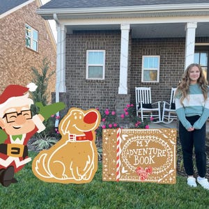May include: A whimsical outdoor Christmas display featuring a cartoon Santa Claus, a dog, and a book with the text "MY ADVENTURE BOOK". The Santa Claus is wearing a red suit and holding a green glove. The dog is tan with a red collar. The book is brown with white lettering.