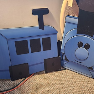 May include: Two blue cardboard train cutouts. One shows the engine with a smiling face and a smokestack. The other shows the train car with three windows and a smokestack. Both have black wheels.