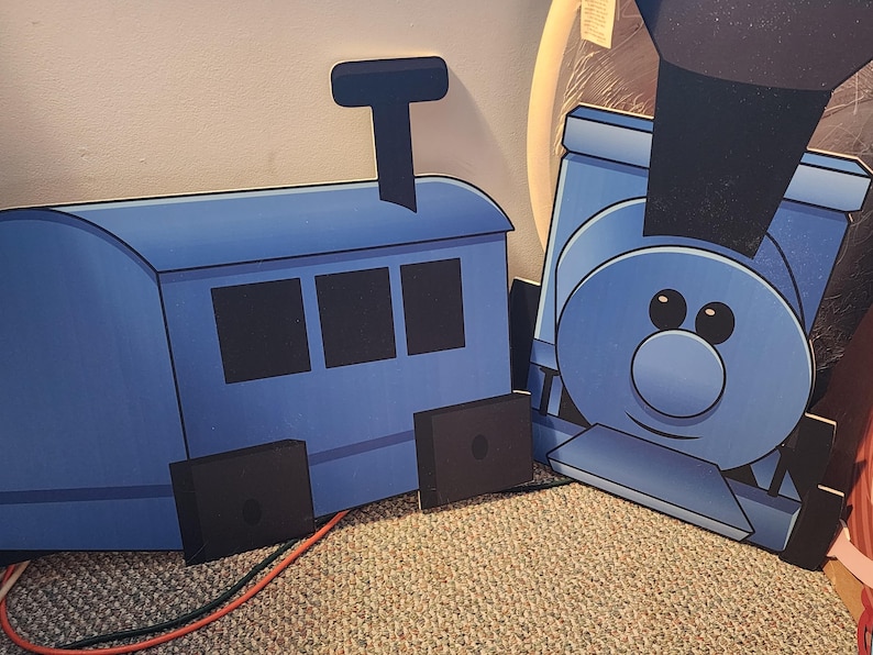 May include: Two blue cardboard train cutouts. One shows the engine with a smiling face and a smokestack. The other shows the train car with three windows and a smokestack. Both have black wheels.