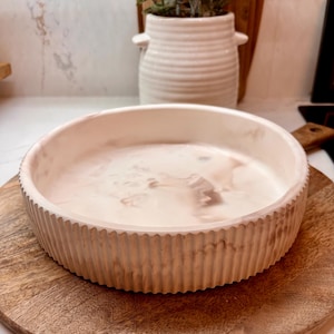 May include: A round, off-white decorative bowl with a ribbed exterior and a marbled interior. The bowl sits on a round wooden serving board. A white ceramic vase with a plant is in the background.