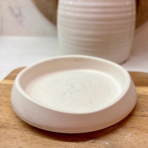 Peut inclure: Un petit bol en céramique blanc cassé avec un bord arrondi, posé sur une planche à découper en bois. Le bol a un design simple et minimaliste. Un pot en céramique blanche avec des poignées est en arrière-plan.