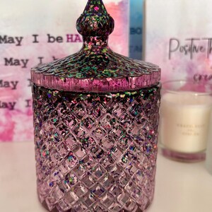 K&ouml;nnte beinhalten: Ein violettes Glas-Kerzengef&auml;&szlig; mit einem Diamantenmuster und einem glitzernden, schillernden Deckel. Das Gef&auml;&szlig; hat ein schwarzes und gr&uuml;nes Glitzerdesign auf dem Deckel. Das Gef&auml;&szlig; steht auf einer wei&szlig;en Oberfl&auml;che.