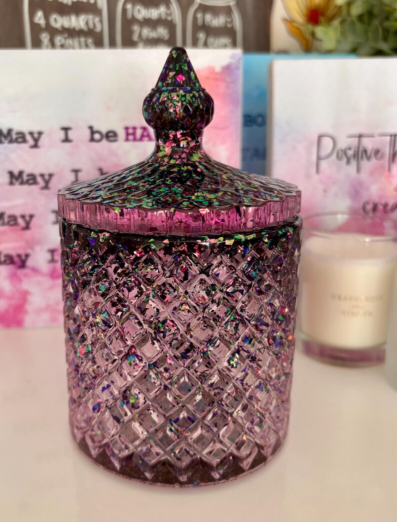 K&ouml;nnte beinhalten: Ein violettes Glas-Kerzengef&auml;&szlig; mit einem Diamantenmuster und einem glitzernden, schillernden Deckel. Das Gef&auml;&szlig; hat ein schwarzes und gr&uuml;nes Glitzerdesign auf dem Deckel. Das Gef&auml;&szlig; steht auf einer wei&szlig;en Oberfl&auml;che.