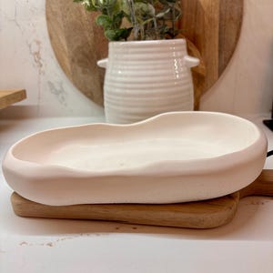 Puede incluir: Una fuente de cerámica rosa claro, de forma oblonga, descansa sobre una pequeña tabla de cortar de madera. Un jarrón de cerámica blanco con asas y una planta están en el fondo. La fuente tiene un diseño liso y curvado.