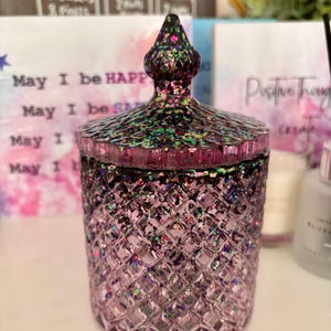 K&ouml;nnte beinhalten: Ein violettes Glas-Kerzengef&auml;&szlig; mit einem Diamantenmuster und einem glitzernden, schillernden Finish. Das Gef&auml;&szlig; hat einen Deckel mit einer spitzen Oberseite. Das Gef&auml;&szlig; steht auf einer wei&szlig;en Oberfl&auml;che.