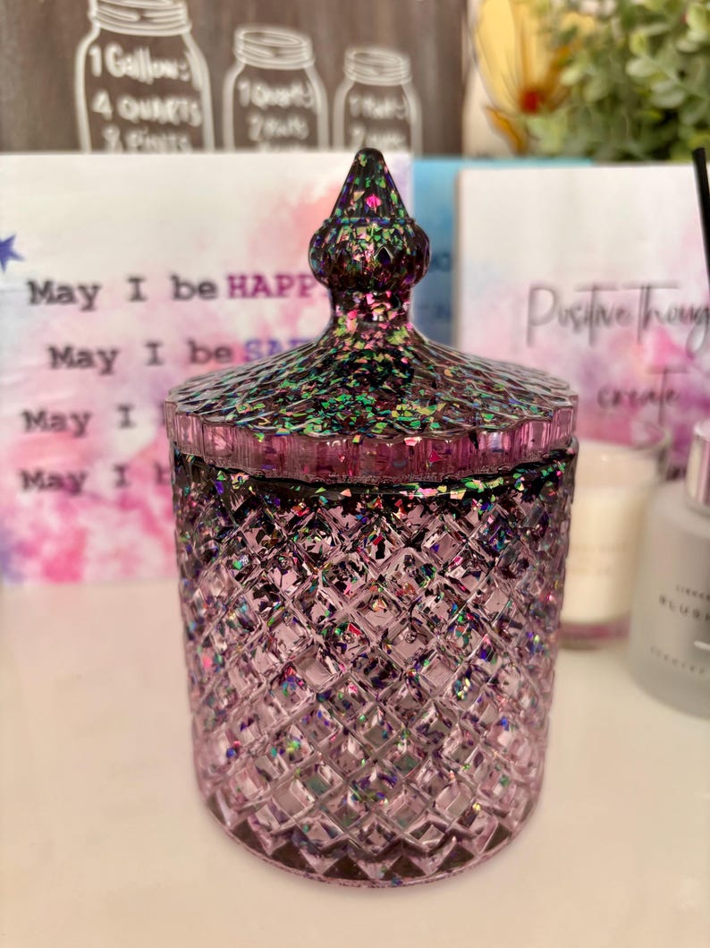 K&ouml;nnte beinhalten: Ein violettes Glas-Kerzengef&auml;&szlig; mit einem Diamantenmuster und einem glitzernden, schillernden Finish. Das Gef&auml;&szlig; hat einen Deckel mit einer spitzen Oberseite. Das Gef&auml;&szlig; steht auf einer wei&szlig;en Oberfl&auml;che.