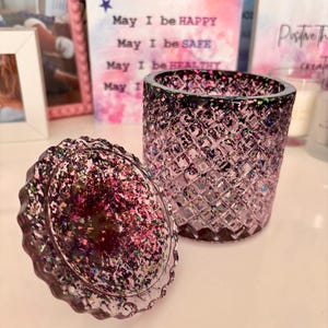 K&ouml;nnte beinhalten: Ein violettes Glas-Kerzengef&auml;&szlig; mit einem Diamantenmuster und einem passenden Deckel. Der Deckel ist mit schimmerndem Glitzer bedeckt. Das Gef&auml;&szlig; steht auf einer wei&szlig;en Oberfl&auml;che.