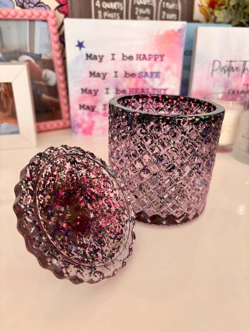 K&ouml;nnte beinhalten: Ein violettes Glas-Kerzengef&auml;&szlig; mit einem Diamantenmuster und einem passenden Deckel. Der Deckel ist mit schimmerndem Glitzer bedeckt. Das Gef&auml;&szlig; steht auf einer wei&szlig;en Oberfl&auml;che.