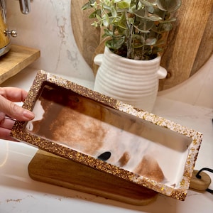 May include: Rectangular decorative tray with a brown and white marbled design. The tray has a gold glitter border. The tray is made of resin and is sitting on a wooden cutting board.