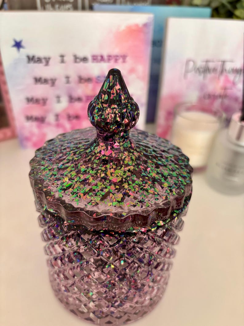 K&ouml;nnte beinhalten: Eine lilafarbene Glasdose mit einem regenbogenfarbenen Glitzerdeckel. Die Dose hat ein Diamantenmuster und der Deckel hat eine spitze Spitze.