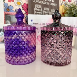 K&ouml;nnte beinhalten: Zwei violette Glaskerzenbeh&auml;lter mit Deckeln. Der Beh&auml;lter links hat ein rosa-violettes Ombre-Design. Der Beh&auml;lter rechts hat eine klare Glasbasis mit schimmerndem Glitzer und einen schwarzen Deckel.