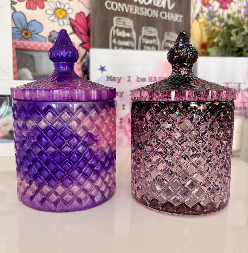 K&ouml;nnte beinhalten: Zwei violette Glaskerzenbeh&auml;lter mit Deckeln. Der Beh&auml;lter links hat ein rosa-violettes Ombre-Design. Der Beh&auml;lter rechts hat eine klare Glasbasis mit schimmerndem Glitzer und einen schwarzen Deckel.