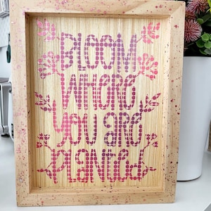 May include: A wooden framed sign with the text "Bloom Where You Are Planted" in pink and purple polka dots. The sign has a light wood background and a pink and white speckled frame.