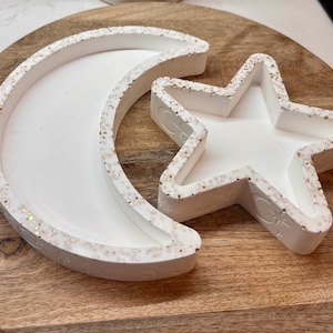 May include: White crescent moon and star-shaped trays with gold glitter trim, set on a wooden board. The trays are likely for decorative or functional use, possibly for jewelry or small items. The design is simple and elegant.