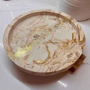 May include: Circular decorative tray featuring a cream and gold marble effect. The tray has a raised rim and is decorated with gold leaf. A small wooden piece is attached to the base. Ideal for displaying items in a home setting.