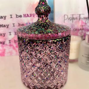 K&ouml;nnte beinhalten: Eine lilafarbene Glasdose mit einem Diamantenmuster und einem glitzernden, schillernden Deckel. Die Dose ist mit einer wei&szlig;en Substanz gef&uuml;llt, m&ouml;glicherweise einer Kerze.