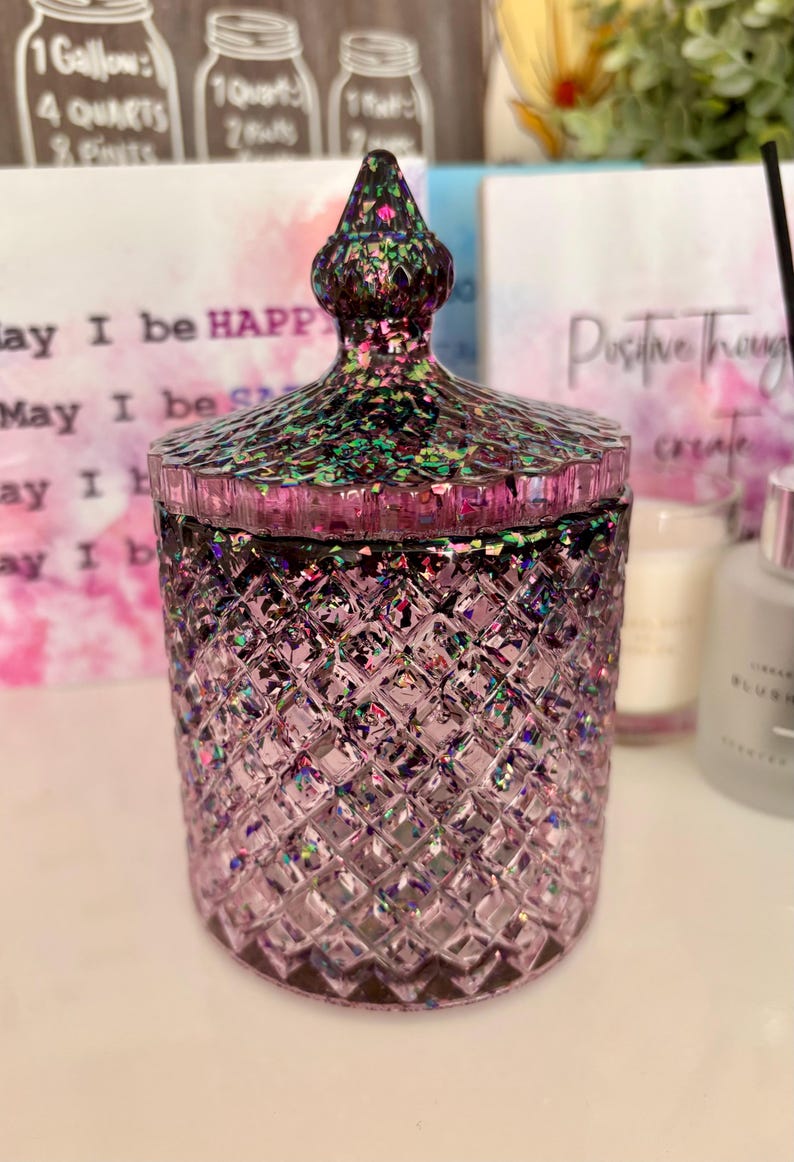 K&ouml;nnte beinhalten: Eine lilafarbene Glasdose mit einem Diamantenmuster und einem glitzernden, schillernden Deckel. Die Dose ist mit einer wei&szlig;en Substanz gef&uuml;llt, m&ouml;glicherweise einer Kerze.
