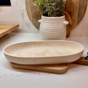 May include: An oval, off-white ceramic serving platter with a speckled texture, on a small wooden chopping board. The platter has a marbled appearance. A white ceramic vase with a plant and a wooden chopping board are in the background.