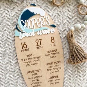 Peut inclure: Une planche de bois en forme de planche de surf avec un motif de vague bleue et blanche. Le texte indique Cooper's first wave, 16 pounds, 27 inches, 8 teeth, ainsi que des surnoms, livres préférés, aliments préférés et étapes de développement.