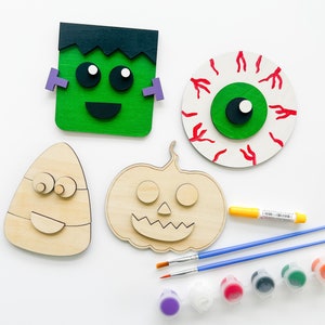 May include: A Halloween craft kit with four unfinished wooden shapes: a pumpkin, a candy corn, a Frankenstein, and an eyeball. The kit includes paint, paintbrushes, and a yellow marker.