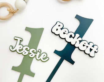 May include: Two green and dark green acrylic cake toppers shaped like the number one with the names "Jessie" and "Beckett" in white lettering.
