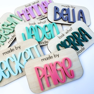 May include: A collection of rectangular wooden plaques featuring raised, colorful acrylic lettering. Each plaque displays the text "made by" or "look what I made" alongside personalized names including Hannah, Bella, Jaden, Corra, Beckett, and Paige.