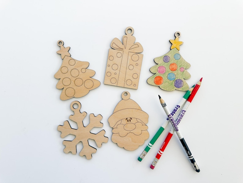 May include: Five unfinished wooden Christmas ornaments: a Christmas tree, a gift box, another Christmas tree, a snowflake, and Santa Claus's face.  The ornaments are light brown and ready to be colored. Three colored pencils—red, green, and black—are also in the image.  The pencils are labeled 'Erasermate.'
