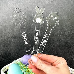 May include: Three clear acrylic Easter egg holders with bunny, chick, and egg designs. Each holder has a name on it: "Emmy", "Emersyn", and "Emmy".