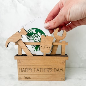 May include: A wooden toolbox with a gift card inside. The toolbox has a hammer, wrench, and saw on the front. The gift card is a Starbucks gift card. The toolbox has the text "HAPPY FATHER'S DAY!" and "love," on the front.