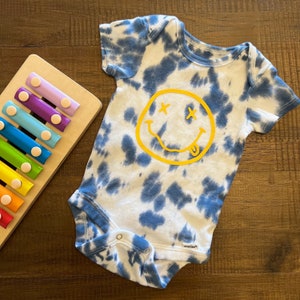 May include: A blue and white tie-dye baby onesie with a yellow smiley face graphic. The smiley face has X's for eyes and a tongue sticking out.