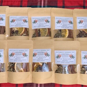 May include: Ten small brown paper bags with clear plastic windows, each containing a mix of dried herbs, spices, and fruit. The bags are labeled "Petunia's Herbal Co. Christmas Morning" and list the ingredients: "Mulled Cinnamon, Clove, Orange, Star Anise".