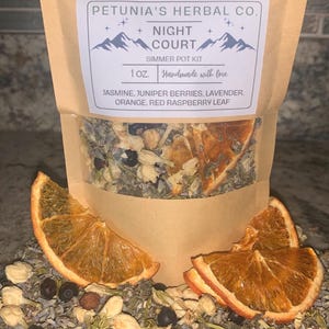 May include: A brown paper bag with a label that reads "PETUNIA'S HERBAL CO. NIGHT COURT SIMMER POT KIT 1OZ. Handmade with love JASMINE, JUNIPER BERRIES, LAVENDER. ORANGE. RED RASPBERRY LEAF". The bag contains a blend of dried herbs and fruit, including orange peels, juniper berries, and lavender.