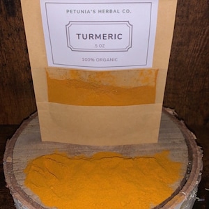 May include: A brown paper pouch with a white label that reads "PETUNIA'S HERBAL CO. TURMERIC 5 OZ 100% ORGANIC". The pouch is filled with bright yellow turmeric powder. The pouch is sitting on a wooden surface.